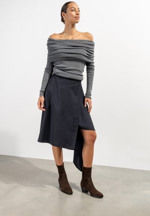 Grijze off-shoulder trui met gerimpelde textuur, gecombineerd met een donkere, asymmetrische pinstripe rok en bruine enkellaarsjes met blokhak.