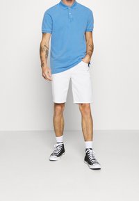 Polo en coton bleu avec un col, associé à un short blanc. Des baskets montantes noires et des chaussettes blanches complètent la tenue.