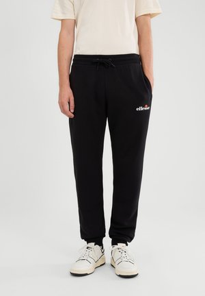 Osoba nosząca czarne spodnie dresowe ellesse ze sznurkiem w talii i białe sneakersy na jednolitym tle.