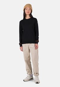 Schwarzer, langärmliger Pullover mit gerippten Bündchen, kombiniert mit cremefarbenen Hosen und grauen Sneakers. Das Model trägt eine beige Strickmütze.