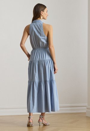 Žena stojaca v interiéri, oblečená v svetlomodrých vrstvených maxi šatách bez rukávov a béžových sandáloch na podpätku, otočená chrbtom k fotoaparátu.