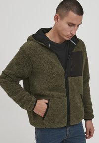 Fuzzy green fleece jacket with a black zippered chest pocket, black accents, and a hood. Front pockets with a textured finish and zipper.