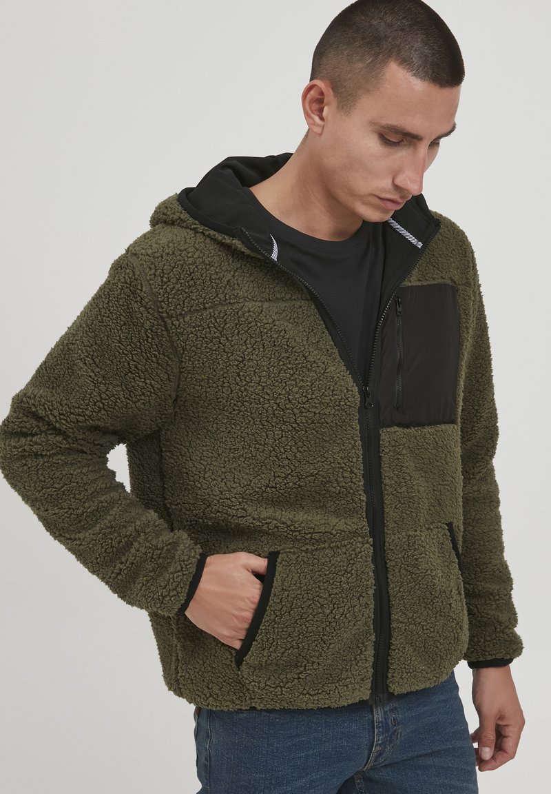 Fuzzy green fleece jacket with a black zippered chest pocket, black accents, and a hood. Front pockets with a textured finish and zipper.