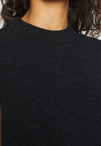 Primo piano di una persona che indossa un maglione senza maniche di colore grigio scuro, con collo a girocollo a coste.