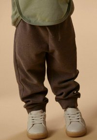Enfant portant un pantalon de survêtement marron et des baskets blanches, debout sur un sol beige avec un haut vert clair partiellement visible.