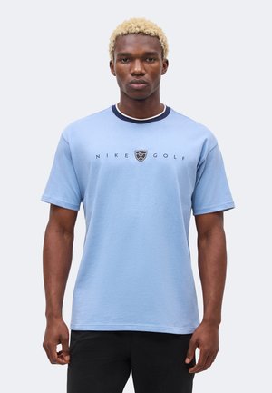 Homme aux cheveux blonds courts portant un t-shirt Nike Golf bleu clair et un pantalon noir, faisant face vers l’avant sur un fond clair uni.