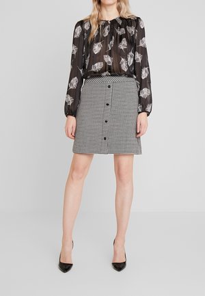 Femme portant un chemisier noir transparent avec des imprimés plumes blanches et une jupe à taille haute noir et blanc à boutons motif pied-de-poule avec des talons noirs.