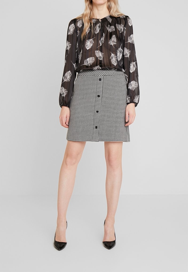 Femme portant un chemisier noir transparent avec des imprimés plumes blanches et une jupe à taille haute noir et blanc à boutons motif pied-de-poule avec des talons noirs.