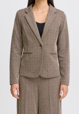 Donna che indossa un blazer marrone a motivo spina di pesce e pantaloni abbinati sopra una maglia nera, in piedi contro uno sfondo chiaro uniforme.