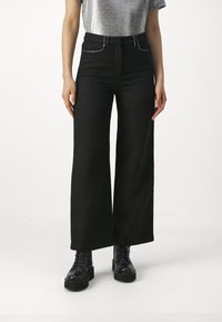 Pantalon large noir en tissu texturé, doté d'une taille haute, de poches avant avec des surpiqûres contrastantes, porté avec des bottes noires à lacets.