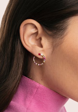 Złota kolczyk kołowy ozdobiony wielobarwnymi kamieniami szlachetnymi, o gładkiej fakturze i zakrzywionym kształcie, noszony na lewym uchu.