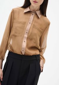 Blouse longue en chiffon marron clair avec un col et des boutons marron plus clair, dotée de deux poches poitrine, associée à un pantalon noir taille haute.