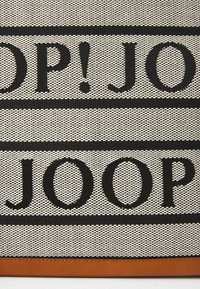 Tapis en tissu tissé noir et crème avec un texte « JOOP! » en gras, accompagné d'une bordure en cuir marron en bas. Texte disposé en bandes horizontales.