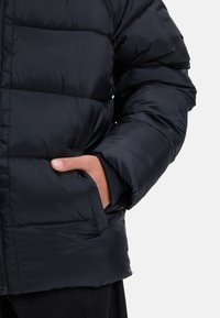 En persons hand delvis inne i den framfickan på en svart pufferjacka med quiltad sömnad och ribbade ärmslut.