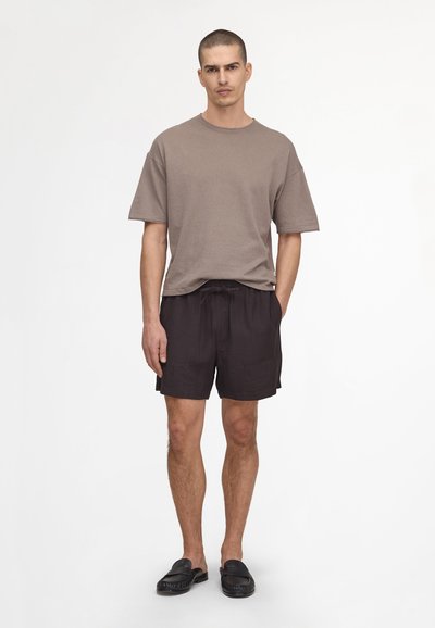 Homme debout, portant un t-shirt oversize manches courtes taupe, un short noir à cordon et des mocassins noirs sans lacets sur fond blanc.