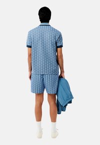 Conjunto de polo y pantalones cortos en azul claro con un patrón geométrico; el tejido parece suave. Una chaqueta azul clara está apoyada sobre el brazo izquierdo.