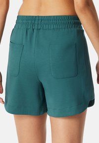 Kvinde iført turkis elastiske shorts med to baglommer, vist fra talje til midt på låret mod en hvid baggrund.