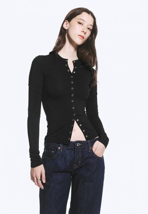 Fiatal nő hosszú barna hajjal, fekete gombos hosszú ujjú felsőt és sötétkék farmert visel, keze a zsebében, sima háttér előtt áll.