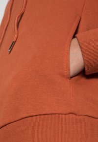 Hand som bär en rostorange sweatshirt med ribbad mudd, som vilar i kengurufickan där snörslutena är synliga.