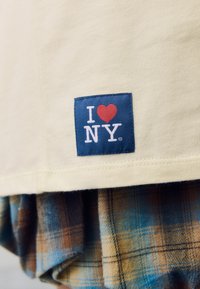 Sárga rövid ujjú ing, amelyen egy kék négyzet alakú folt látható fehér és piros "I ♥ NY" felirattal. Rétegezve kék, barna és bézs kockás anyaggal.