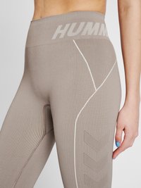 Leggings a coste beige con alta vita featuring il logo "HUMM" in grigio chiaro. Il dettaglio laterale include cuciture bianche e accenti a zigzag.