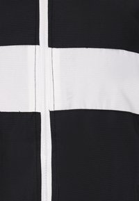 Giacca con zip in bianco e nero, caratterizzata da un tessuto testurizzato con una larga striscia bianca che attraversa il petto, design strutturato e pannelli a contrasto.