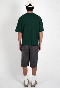 Muž nosi tmavo zelené oversized tričko, sivé nohavice po kolená s prúžkami, biele ponožky, béžové sandále a béžovú čiapku, stojí pred jednoduchým pozadím.