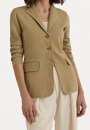 Frau trägt ein beige geknöpftes Blazer mit Klappentaschen und langen Ärmeln über einem cremefarbenen Top und cremefarbenen plissierten Hosen.
