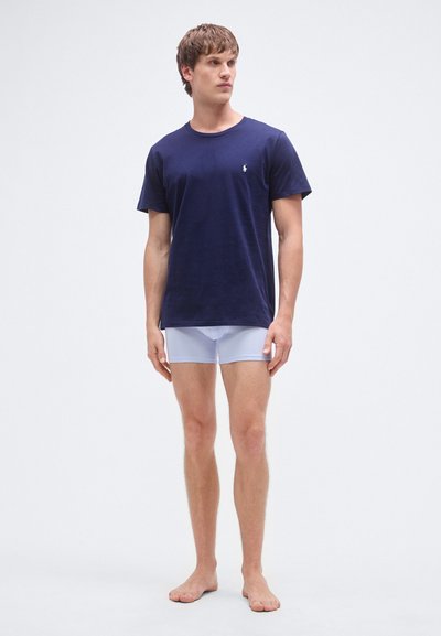 T-shirt azul-marinho em algodão com decote redondo e mangas curtas, combinada com boxers azul claro. Modelo está descalço contra um fundo simples.