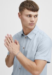 Lichtblauw kortemouwen shirt met een button-down kraag en houten knopen. De handen van het model zijn samengevoegd, met een zilveren ketting armband.