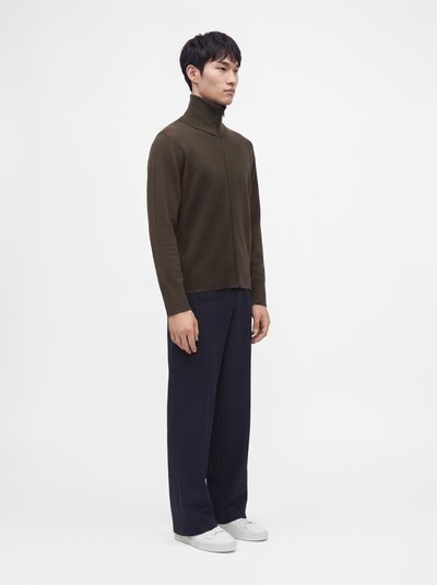 Jeune homme debout portant un pull zippé col montant vert foncé, un pantalon large bleu marine et des baskets blanches sur un fond uni.