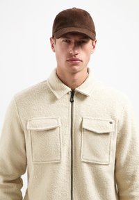 Beige fleecjas met een ritssluiting aan de voorkant, twee borstzakken en een kraag. Bruine pet met een ronde kroon en een platte rand.