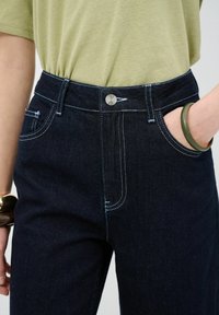 Jeans in denim blu scuro con cuciture a contrasto, chiusura con bottone in metallo e tasche frontali. Abbinati a una t-shirt in cotone verde chiaro.