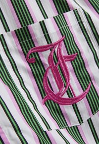 Chemise en tissu rayé présentant des lignes verticales en rose, vert et noir avec une lettre "T" rose brodée en évidence sur la poche.