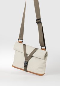 Beige Umhängetasche mit braunem verstellbarem Riemen, Rollverschluss, schwarzer Kunststoffschnalle und braunem Lederbesatz am unteren Rand.