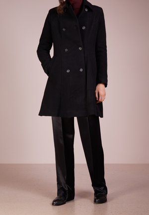 Persona que lleva un abrigo negro de doble botonadura, pantalones negros, zapatos negros y un suéter de cuello alto rojo oscuro con una mano en el bolsillo.