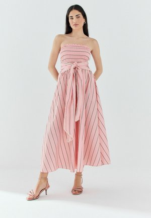 Frau steht und trägt ein trägerloses pinkfarbenes Kleid mit schwarzen vertikalen Streifen und einer großen gebundenen Schleife in der Taille, kombiniert mit pinkfarbenen Sandalen mit Absatz.