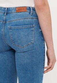 Haalar Blue denim -vaatteet, joissa on keskikorkea istuvuus, ruskea nahkainen "ONLY" -logotarran, kaksi takataskua ja näkyvissä olevat ompeluyksityiskohdat.