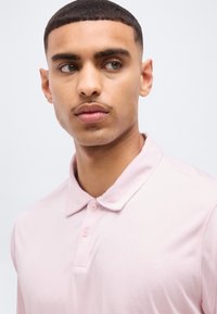 Νέος άνδρας με κοντά σκούρα μαλλιά και μουστάκι, ντυμένος με ανοιχτό ροζ πόλο πουκάμισο με γιακά, κοιτάζοντας ελαφρώς προς το πλάι.