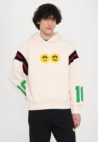 Cream pullover hoodie with black and red stripe details on sleeves, featuring yellow smiley graphics and a front pocket; paired with black pants.