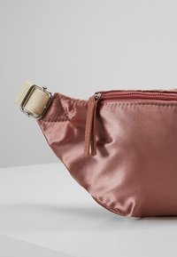 Close-up van een stoffige roze satijnen heuptas met een ritssluiting en een beige verstelbare band met een zilveren gesp op een witte ondergrond.