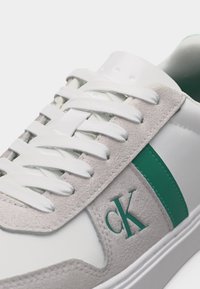 Λευκό αθλητικό παπούτσι με πάνελ από ανοιχτό γκρι σουέντ, τιρκουάζ ρίγα, λευκά κορδόνια και τιρκουάζ λογότυπο "cK" στο πλάι κοντά στα κορδόνια.