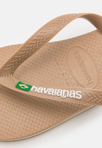 Tan spalvota guma šlepetė su tekstūruotu padu, su įstrižiniu raštu ant dirželių ir logotipu su Brazilijos vėliavos akcentu.