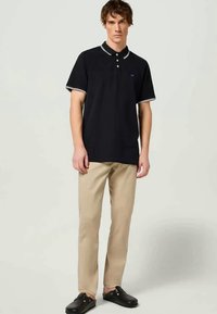 Camisa polo azul con cuello y bordes de manga en blanco, combinada con pantalones beige y zapatos negros de deslizarse, de pie contra un fondo neutro.