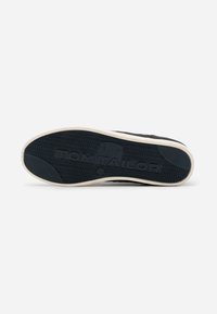Semelle de sneaker noire avec un motif texturé, bord en caoutchouc beige clair, et des détails de logo en relief, présentant un design incurvé pour un meilleur soutien.