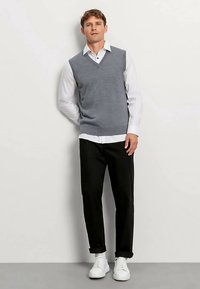 Grijs gebreid v-halsvest over een wit shirt, gecombineerd met zwarte broek met opgerolde pijpen en witte sneakers, tegen een lichte achtergrond.