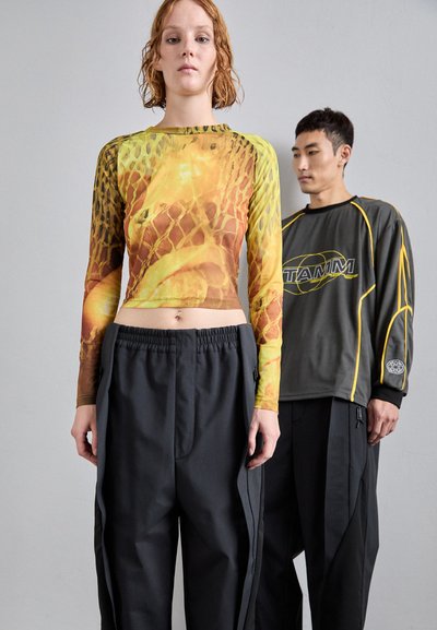 Długi rękaw z żółtym i pomarańczowym wzorem węża; czarne, luźne spodnie z elastycznym pasem i bocznymi kieszeniami na zamek. Męska figura w szarym koszuli w tle.