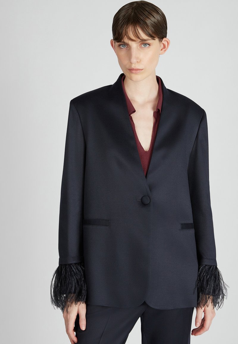 Granatowa marynarka o gładkiej fakturze, z pojedynczym zapięciem na guzik i czarnymi piórami przy mankietach. Posiada dwie przednie kieszenie.