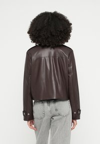 Brūna ādas saīsināta jaka ar gludu tekstūru, plecu epauletēm un sprādzēm, kopā ar pelēnām augsta vidukļa džinsām.