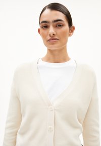 Cremfarbener Cardigan aus strukturiertem Strickmaterial, mit V-Ausschnitt, langen Ärmeln und zwei Knopfverschlüssen vorne. Über einem weißen Hemd getragen.